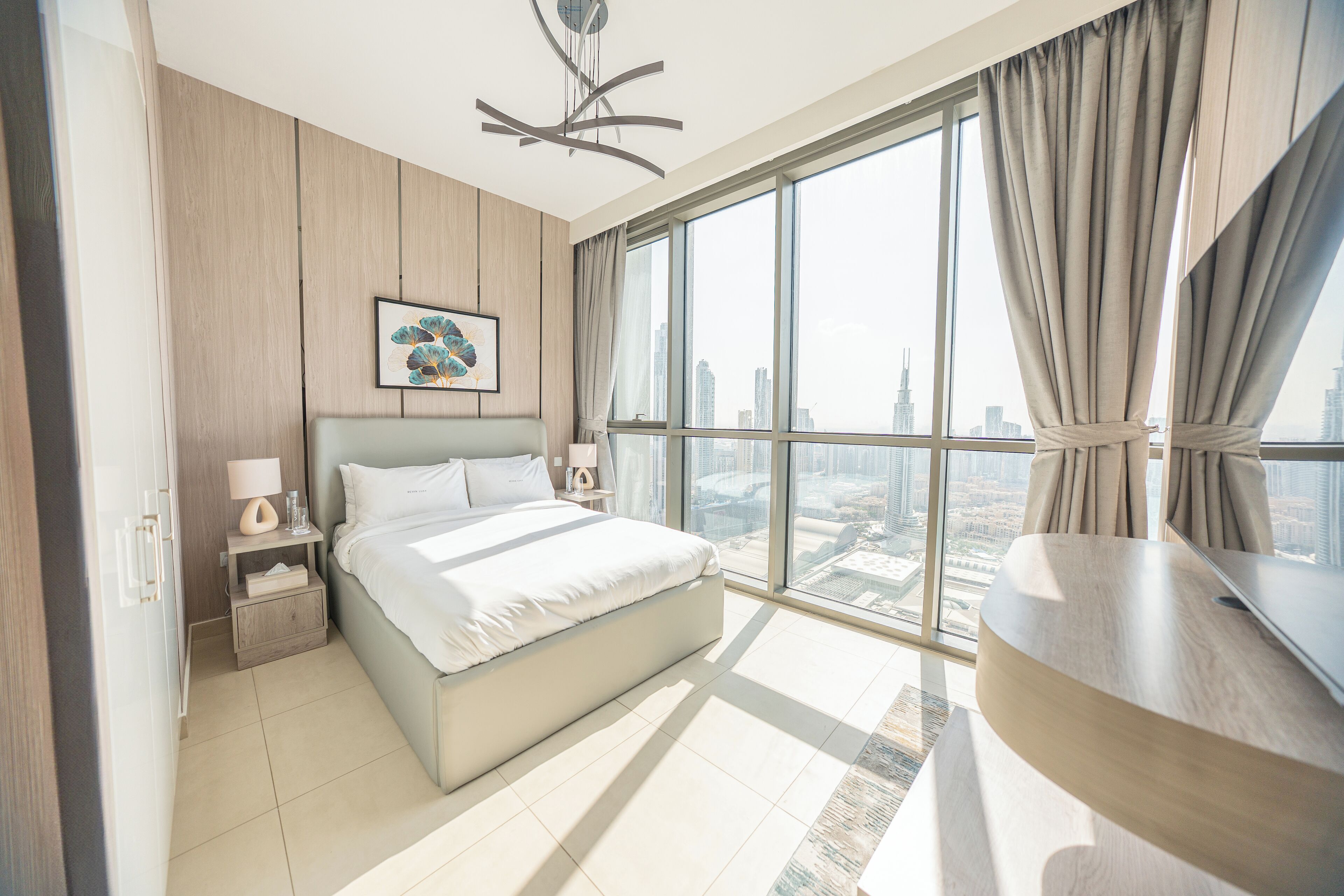Un dormitorio moderno con una cama grande, una mesita de noche y vistas a un paisaje urbano a través de amplios ventanales.