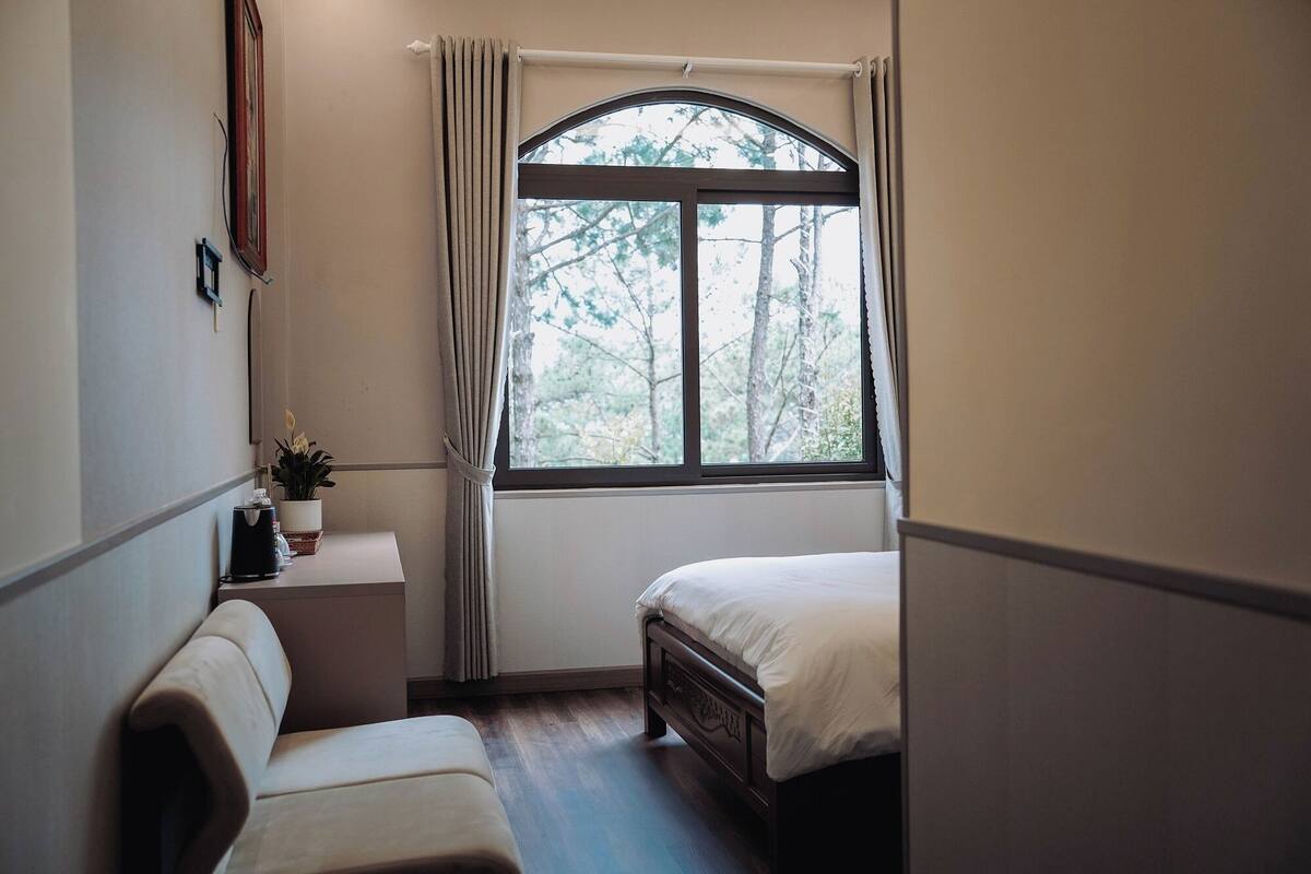 Une chambre à coucher avec un lit, une table de chevet, une petite table avec une plante et une fenêtre avec des rideaux.