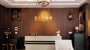 Zona de recepción de hotel con mostrador, señalización, un reloj, una araña y un área de estar con sofá y sillones.