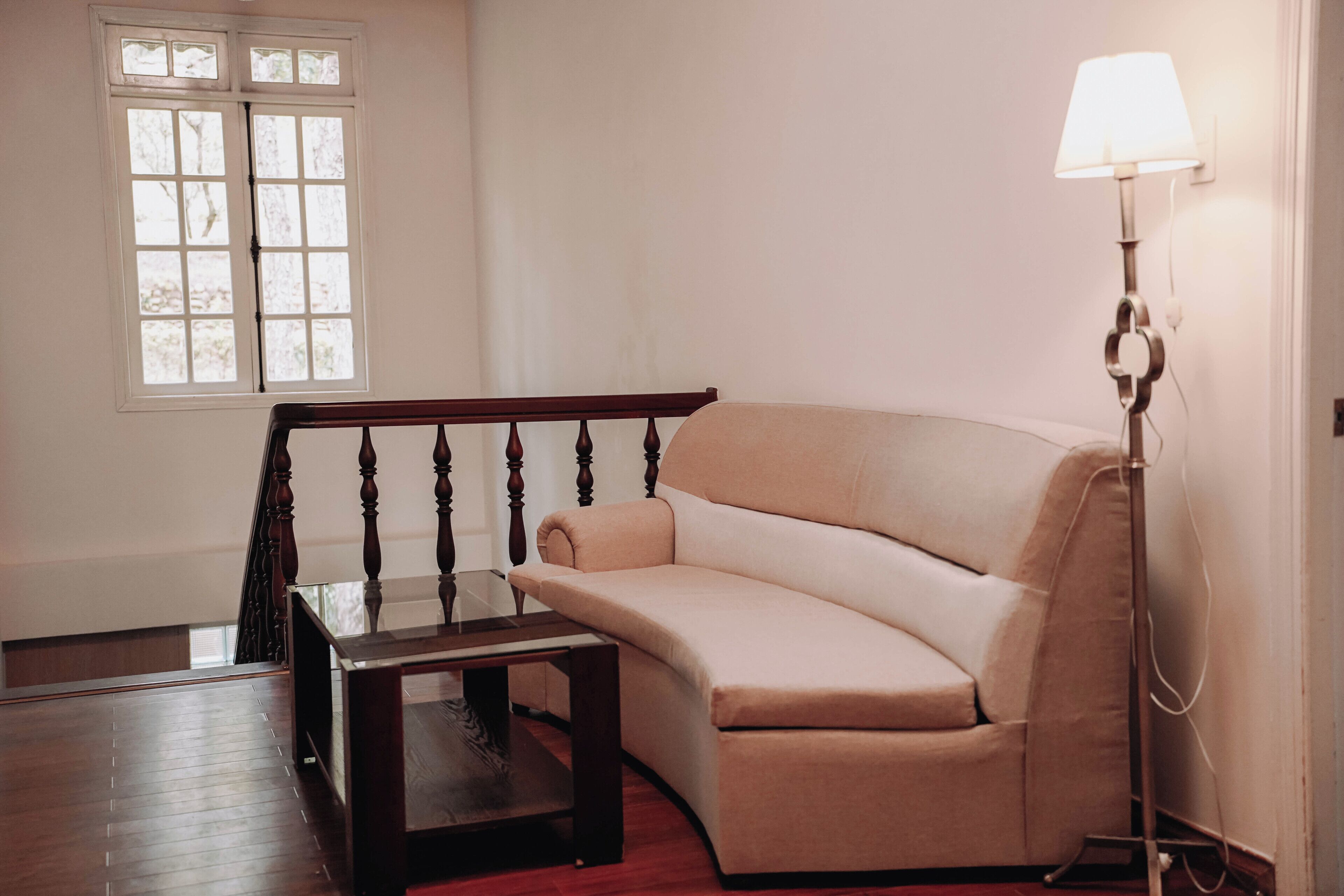A room with a curved sofa, a glass coffee table, a wooden floor, a staircase with a wooden railing, and a floor lamp.