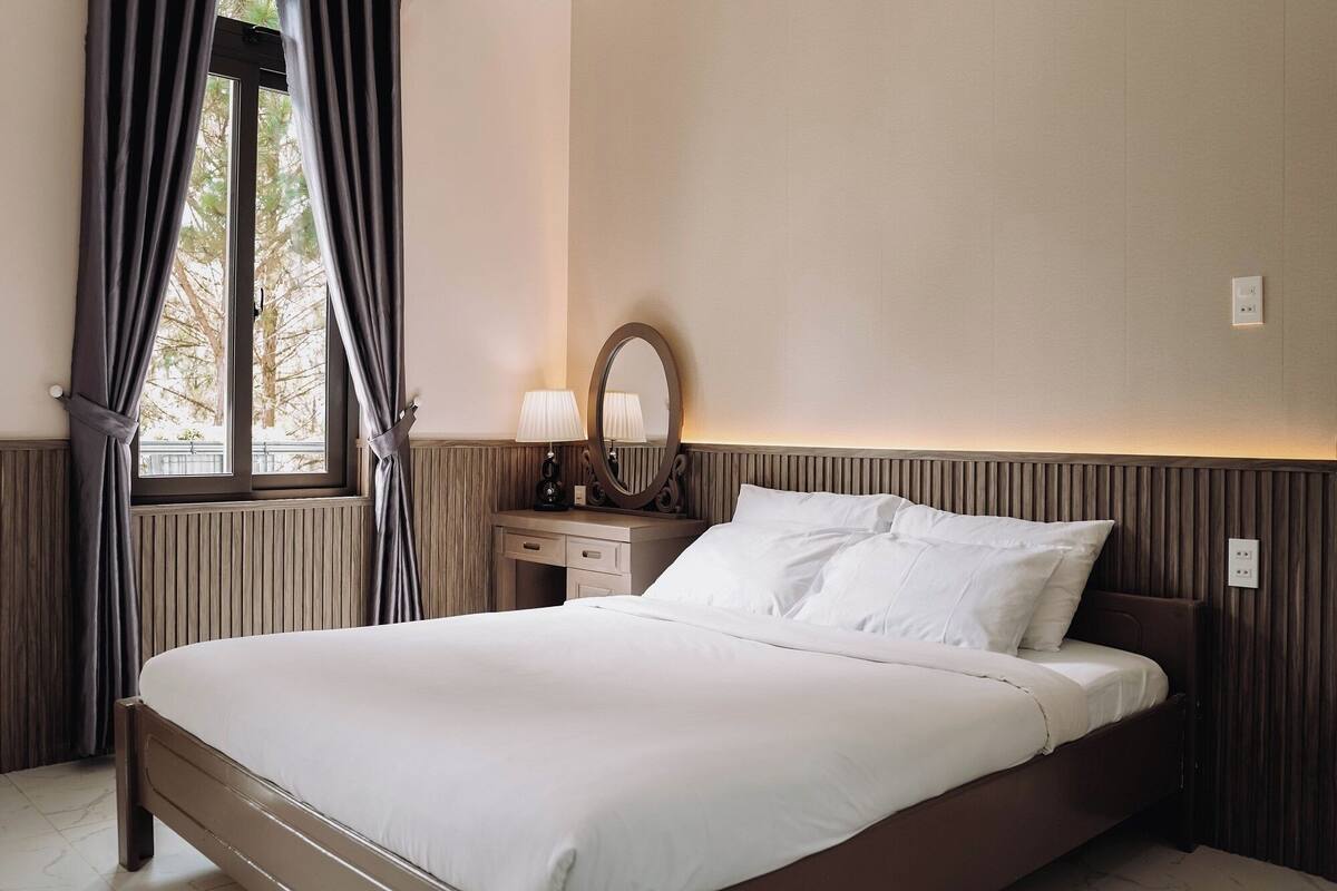 Une chambre à coucher avec un lit, une table de chevet, une lampe, un miroir et une fenêtre avec des rideaux.