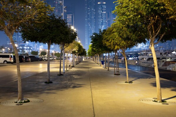 A nighttime urban waterfront with a tree-lined promenade, high-rise buildings in the background, and boats docked along the water. - Luxury Branded Suite with Beach Access & Sea Views (Dubai)
