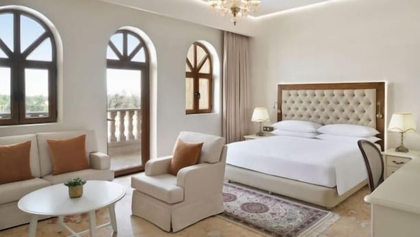 A hotel room with a large bed, a sofa, a chair, a small table, and large windows. - Al Samriya Doha Autograph Collection (Ash Shahaniya)