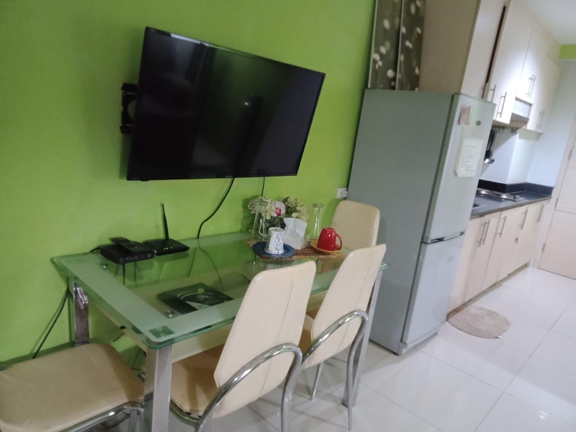 A dining area with a glass table, four chairs, a TV mounted on the wall, and a refrigerator in the background.