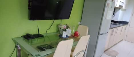 Un comedor con una mesa de vidrio, cuatro sillas, un televisor montado en la pared y un refrigerador al fondo.