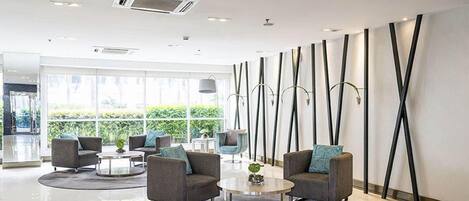 Een moderne lobby met grijze banken, een ronde salontafel en een groot raam met uitzicht op groen.
