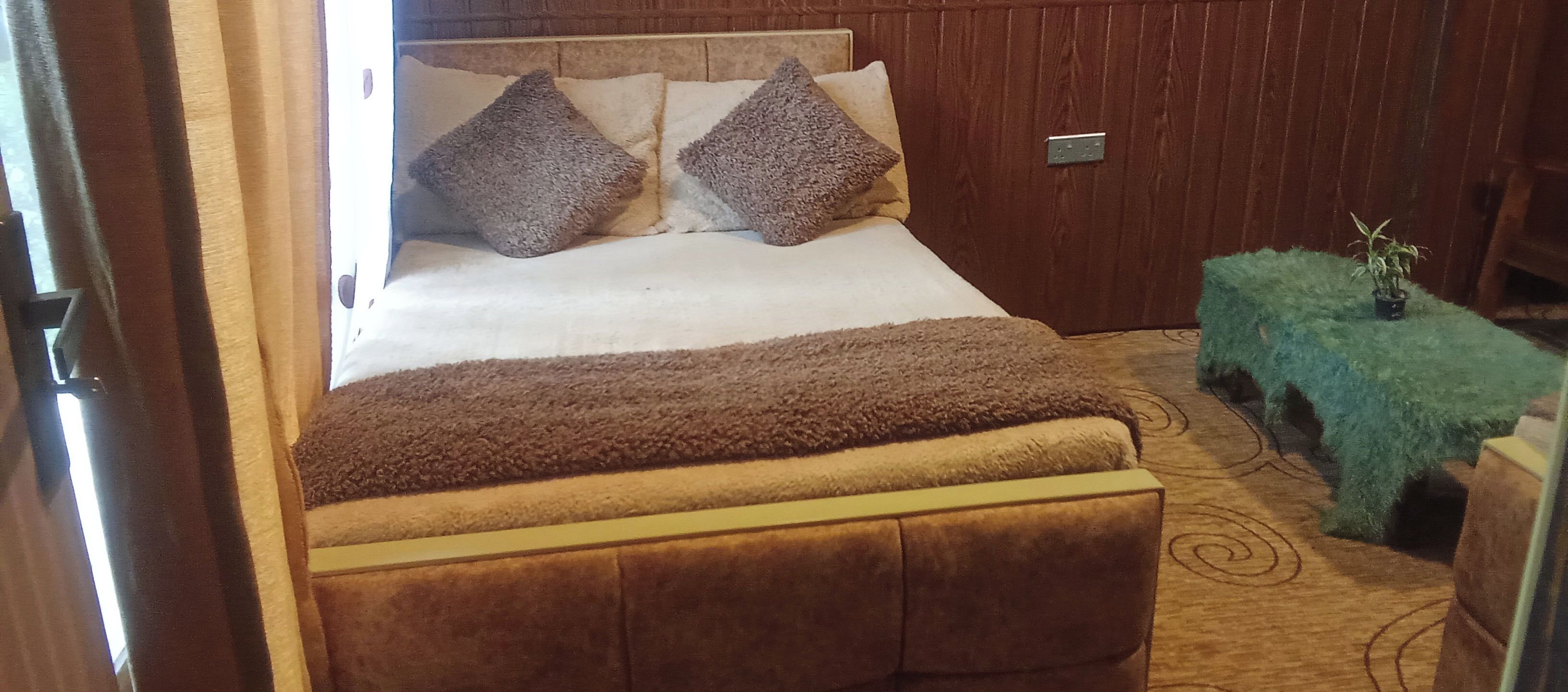 Ein Bett mit Kopfteil, zwei Kissen, einer braunen Decke und einem grünen Nachttisch mit Pflanze.