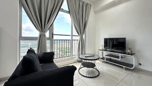 Een moderne woonkamer met een zwarte bank, een televisie op een witte dressoir, een kleine ronde tafel en grote ramen met gordijnen.