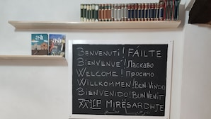 A room with a white table and four black chairs, a grey sofa, and a bookshelf with books. A blackboard with multilingual welcome messages is on the wall. - Casa del Prof - Holiday apartment in the historic center (Asti)