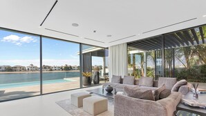 Ein modernes Wohnzimmer mit einer großen Glaswand, die auf einen Pool und ein Gewässer blickt. In der Ferne sind Gebäude zu sehen.