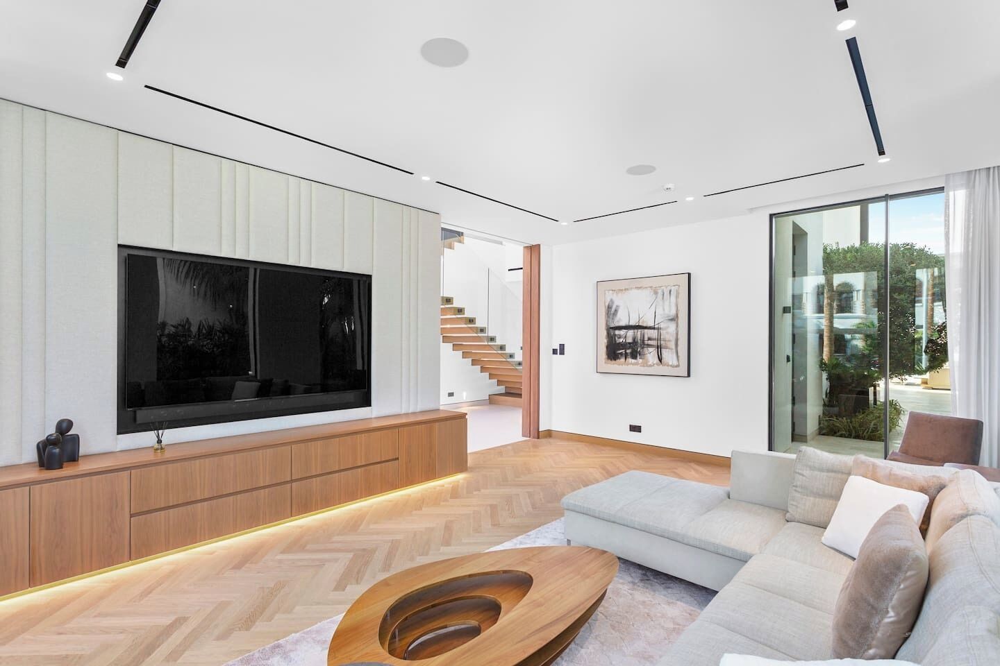 Un salon moderne doté d’un grand téléviseur à écran plat, d’une console en bois, d’un parquet à motifs en chevrons, d’un canapé gris et d’une porte en verre donnant sur un escalier.
