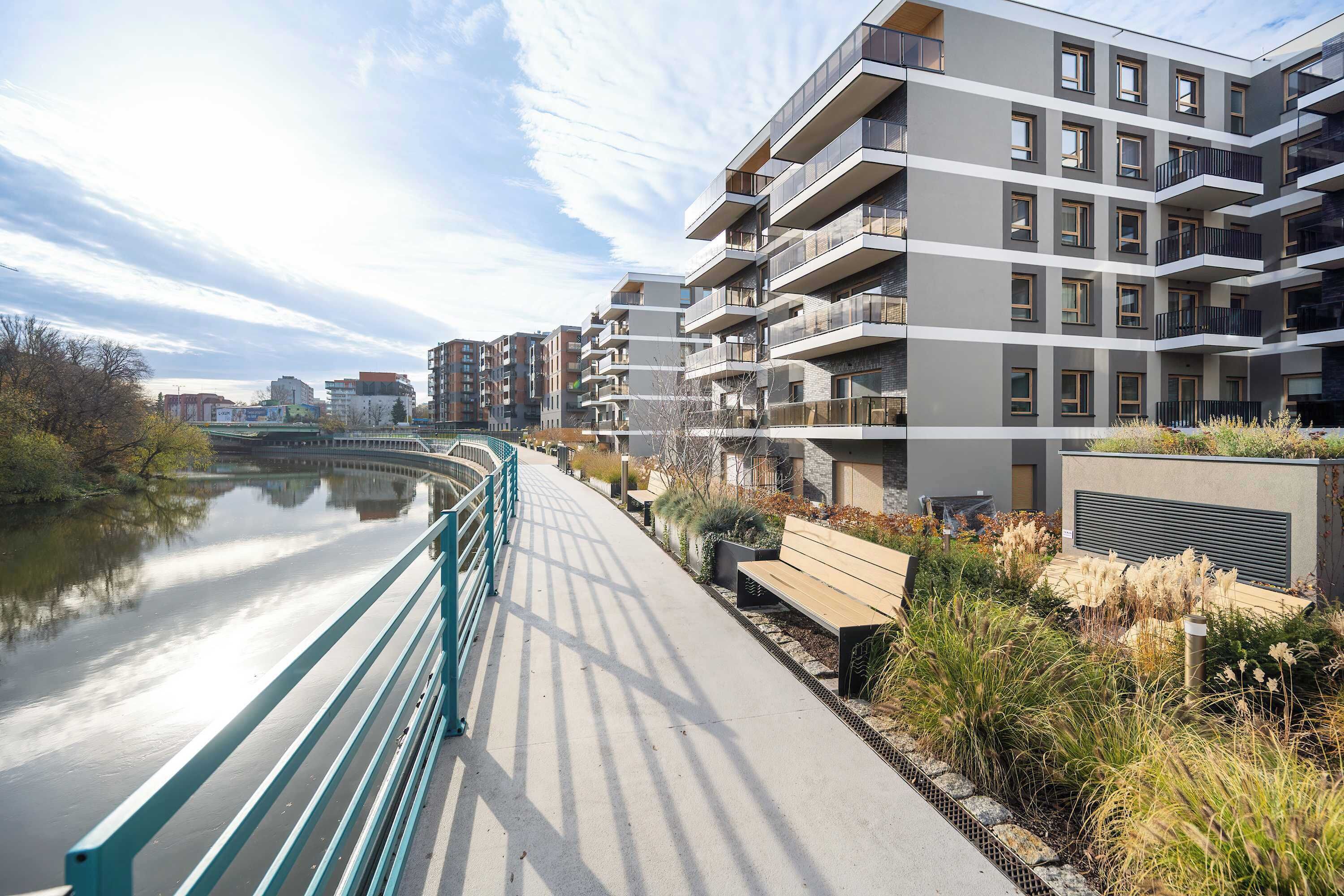 A modern apartment building with balconies, a river in the foreground, and a pedestrian bridge.