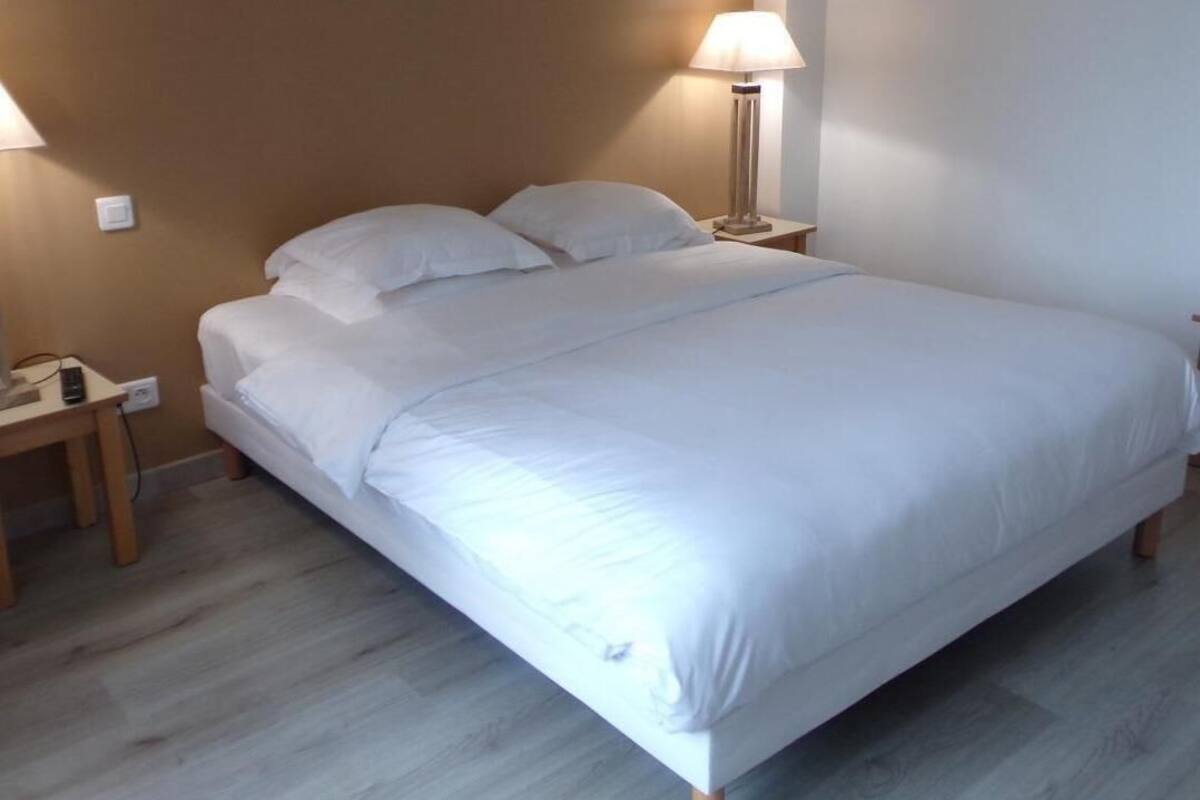Ein ordentlich bezogenes Bett mit weißen Leinen, ein Nachttisch mit Lampe und eine Steckdose an der Wand.