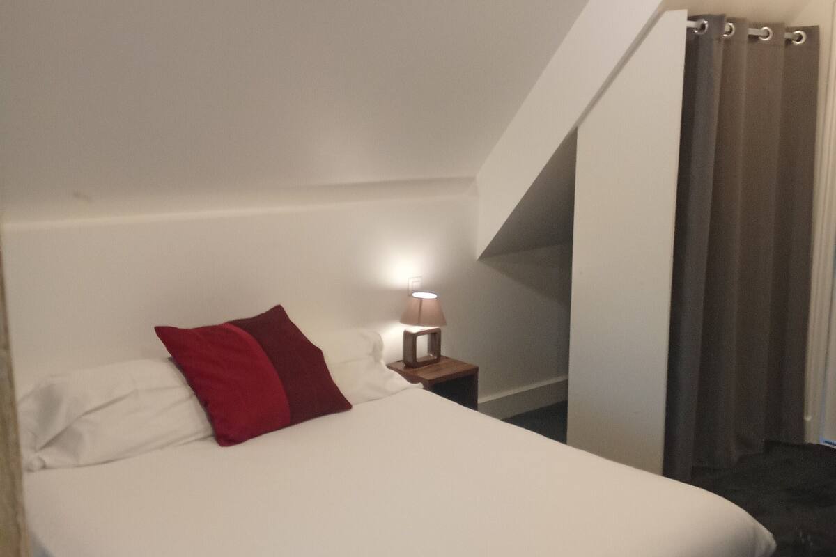 Ein Schlafzimmer mit einem Bett, einem Nachttisch mit Lampe und einem Fenster mit Vorhängen.