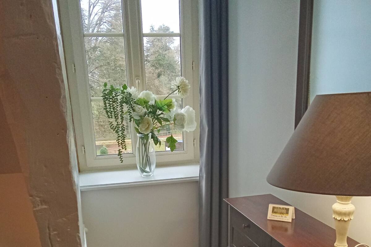 Ein Zimmer mit Schreibtisch, Stuhl, Lampe, Blumen in einer Vase und einem Vorhangfenster.