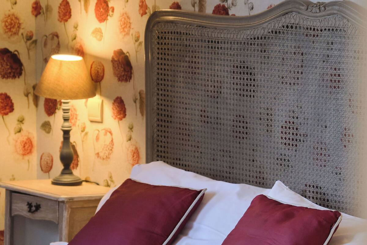 Ein ordentlich bezogenes Bett mit zwei roten Kissen und einer weißen Bettdecke, eine Lampe auf einem Nachttisch und ein floraler Wandbehang.