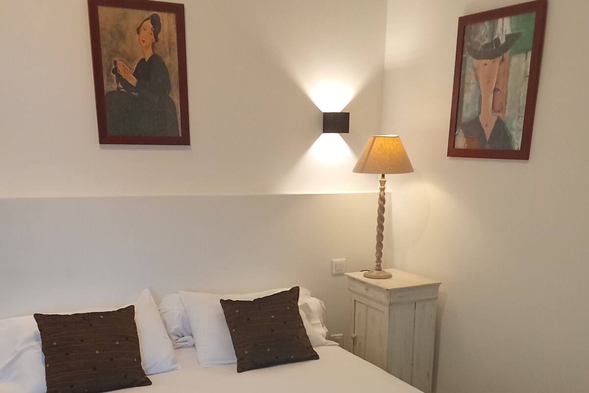 Ein ordentlich gemachtes Bett mit weißen und dunkelbraunen Kissen, einer Nachttischlampe sowie zwei gerahmten Kunstwerken an der Wand.
