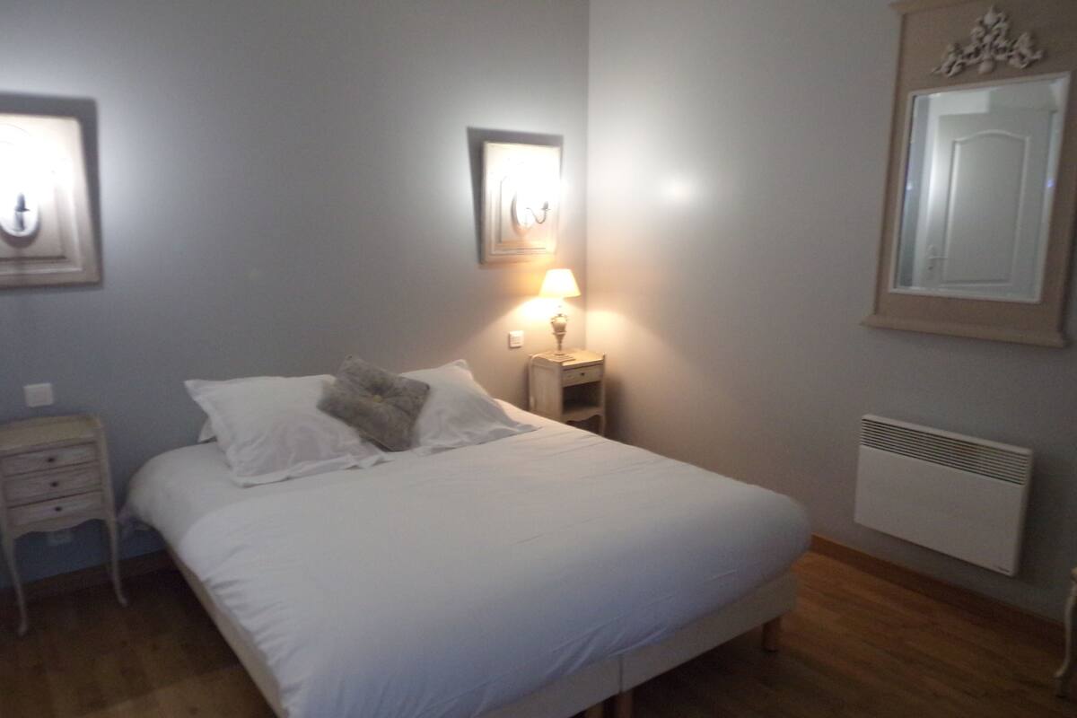 Ein ordentlich bezogenes Bett mit weißen Leinentüchern und einem grauen Kopfkissen, ein Nachttisch mit einer Lampe sowie ein an der Wand befestigter Spiegel.