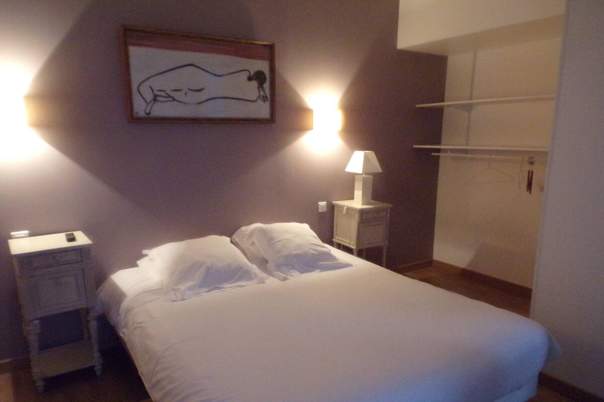 Ein ordentlich bezogenes Bett mit weißen Leinen, ein Nachttisch mit Lampe und ein gerahmtes Bild an der Wand.