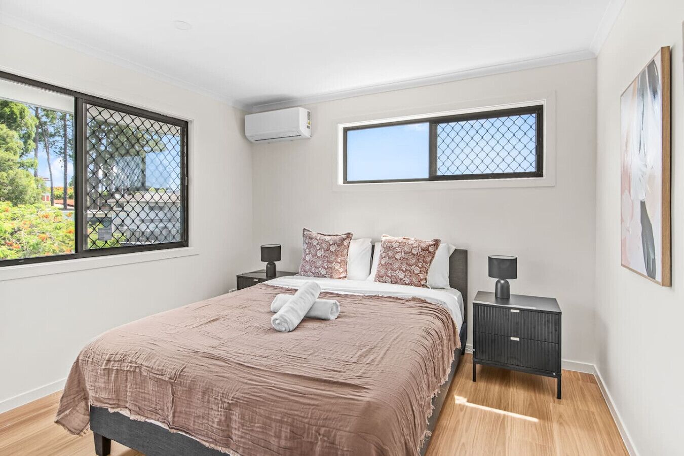 Un dormitorio con cama, dos mesitas de noche, un equipo de aire acondicionado y dos ventanas con mosquiteras.
