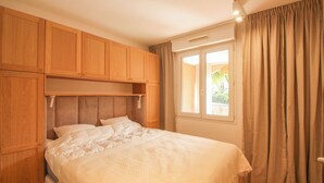 A bedroom with a bed, wooden cabinets, a window with curtains, and a ceiling vent. - Cosy T3 5 minutes from beaches and town center (Sainte-Maxime)