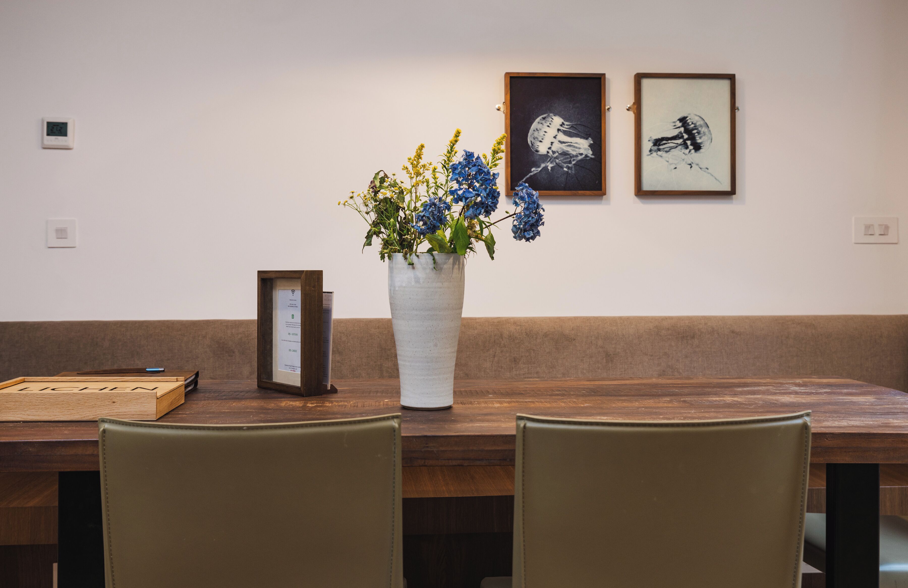 Une table en bois sur laquelle est posé un vase blanc rempli de fleurs, une œuvre d’art encadrée au mur et une boîte en bois portant le mot « LICHEN ».