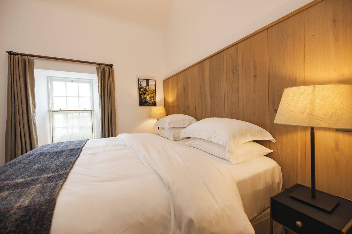 A hotel room with a wooden headboard, a bed with white linens, a bedside lamp, and a window with curtains.
