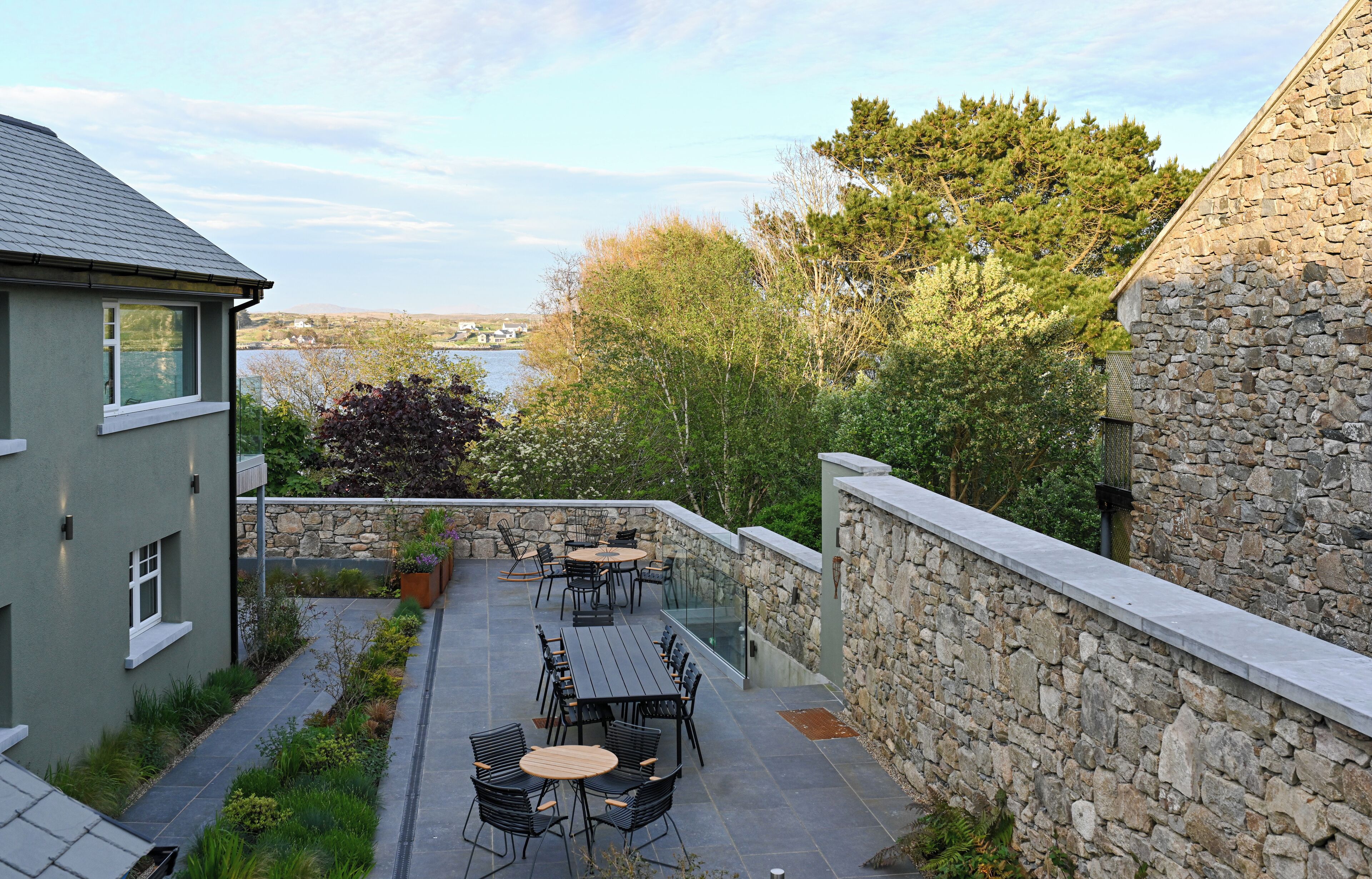 Eine Terrasse mit Sitzgelegenheiten, eine Steinmauer und Blick auf ein Gewässer und Bäume.