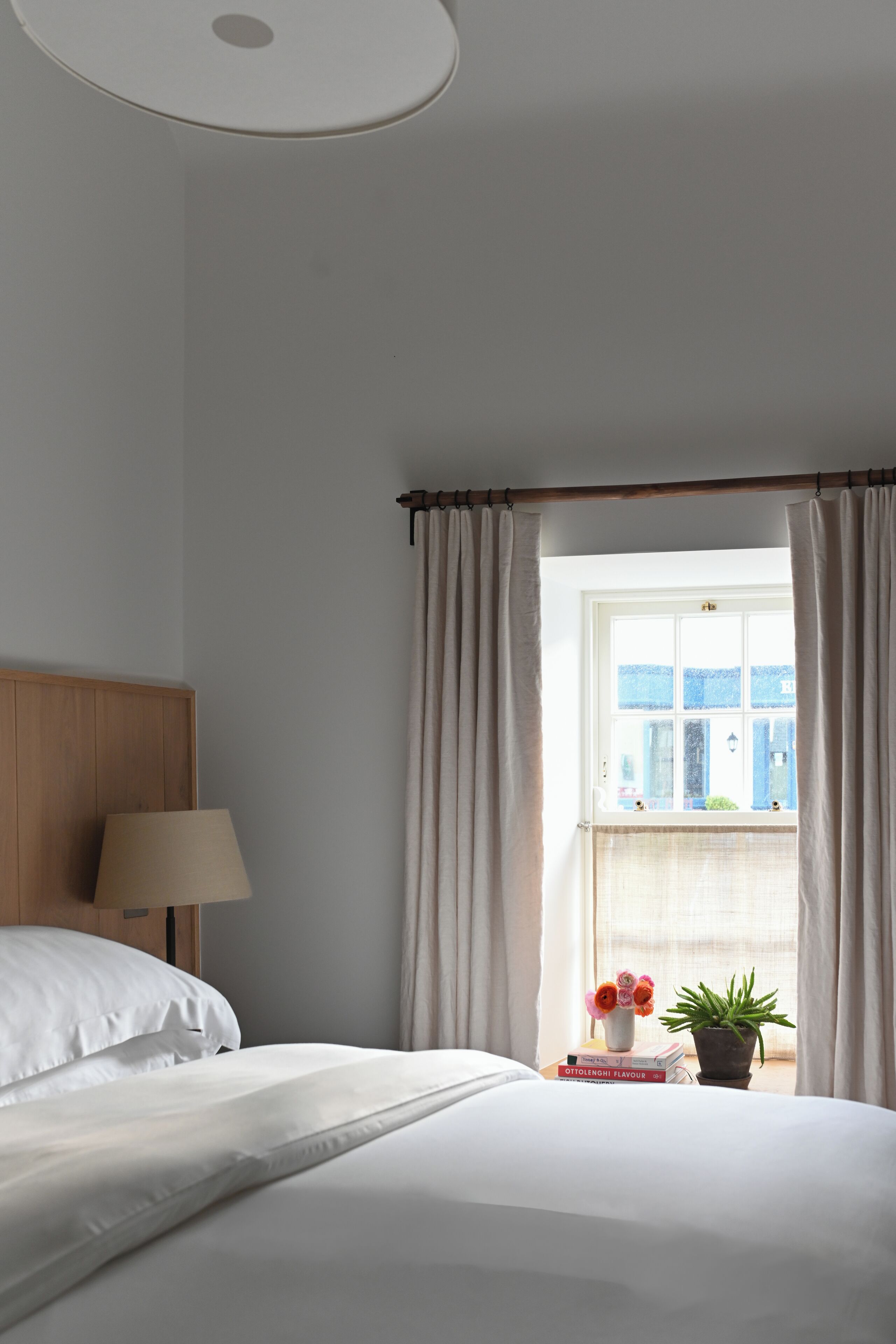 A neatly made bed with white linens, a bedside lamp, a window with curtains, a small table with a vase of flowers, and a potted plant.