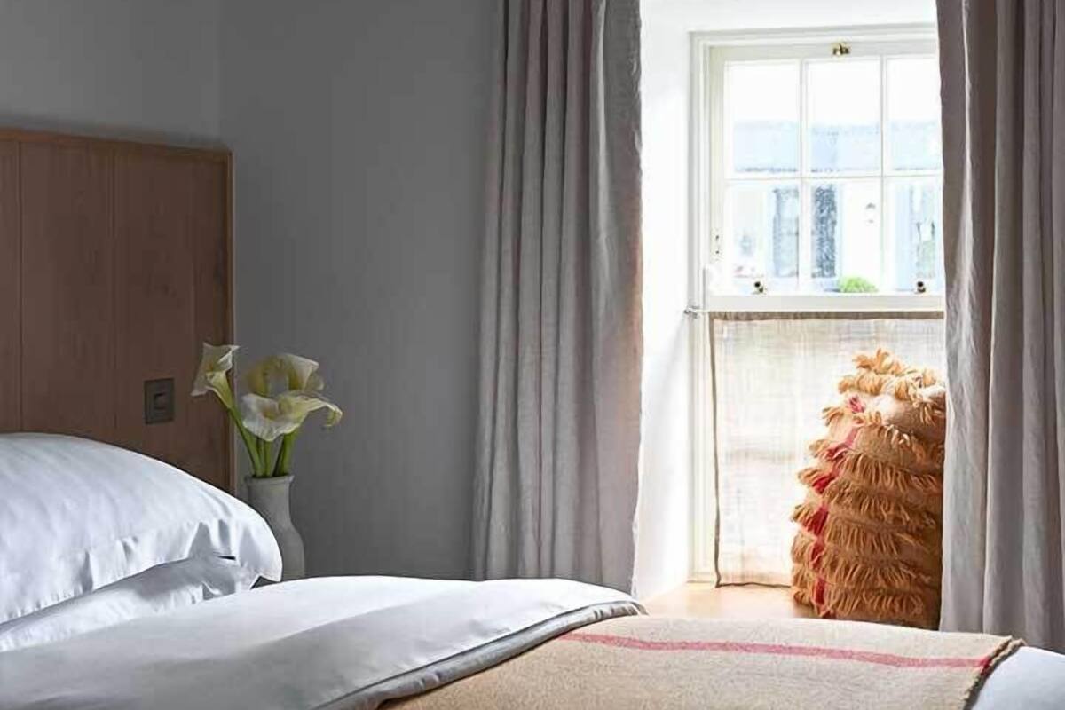 A bedroom with a bed, a window with curtains, a vase with flowers, and a view of the outside.