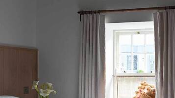 A bedroom with a bed, a window with curtains, a vase with flowers, and a view of the outside.