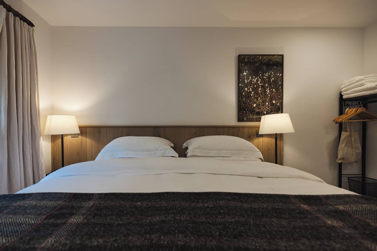 A hotel room with a large bed, two bedside lamps, a window with curtains, a wall-mounted artwork, and a towel rack with towels.