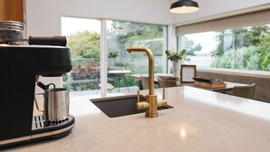 A modern kitchen with a coffee machine, a gold faucet, and a view of the outdoors through large windows. - Within the Village (Roundstone)