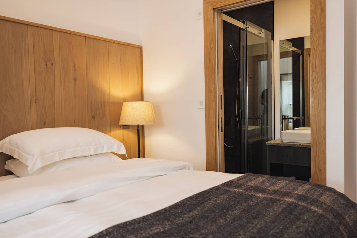 A hotel room with a wooden headboard, a bed with white linens, a floor lamp, and a glass shower door.