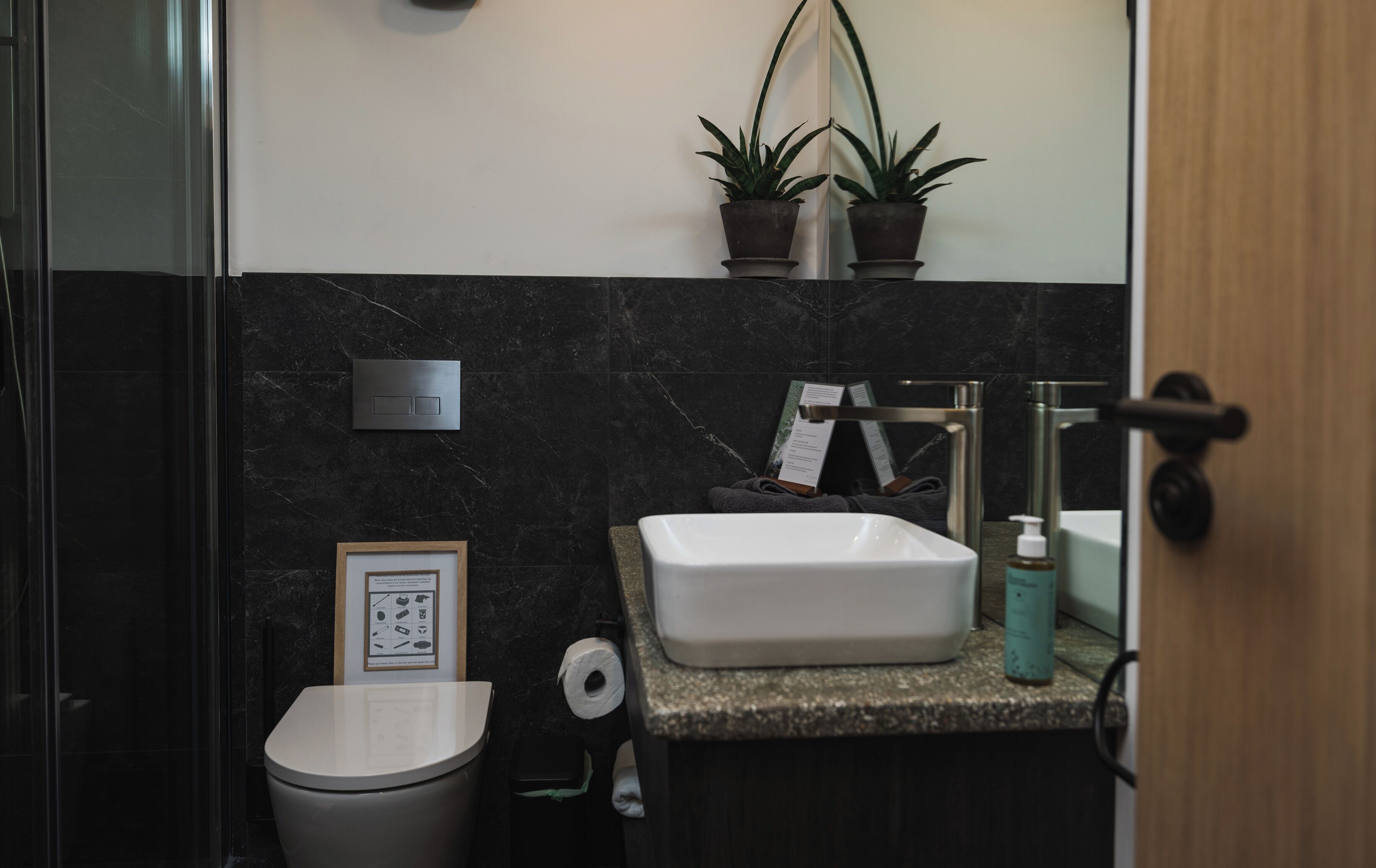 A bathroom with a white sink, dark marble walls, a framed informational poster, a toilet, and two potted plants.