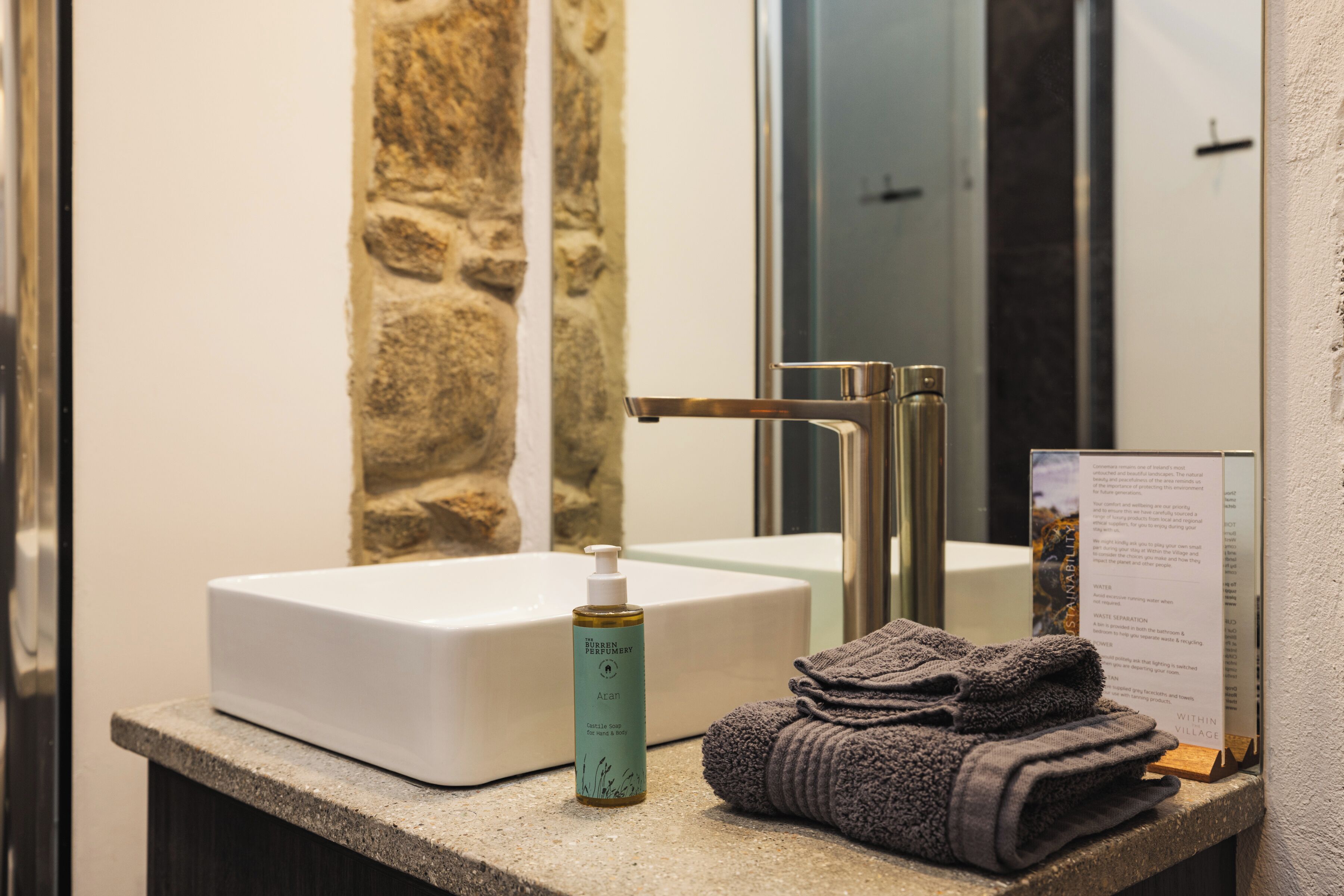 A bathroom with a white sink, a bottle of Aran body wash, and a folded towel on a stone countertop.