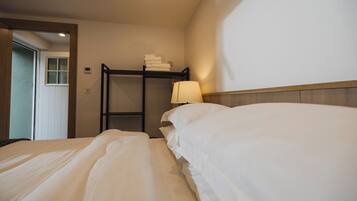 A hotel room with two beds, a wooden bunk bed with towels, a bedside lamp, and a door leading to another room.