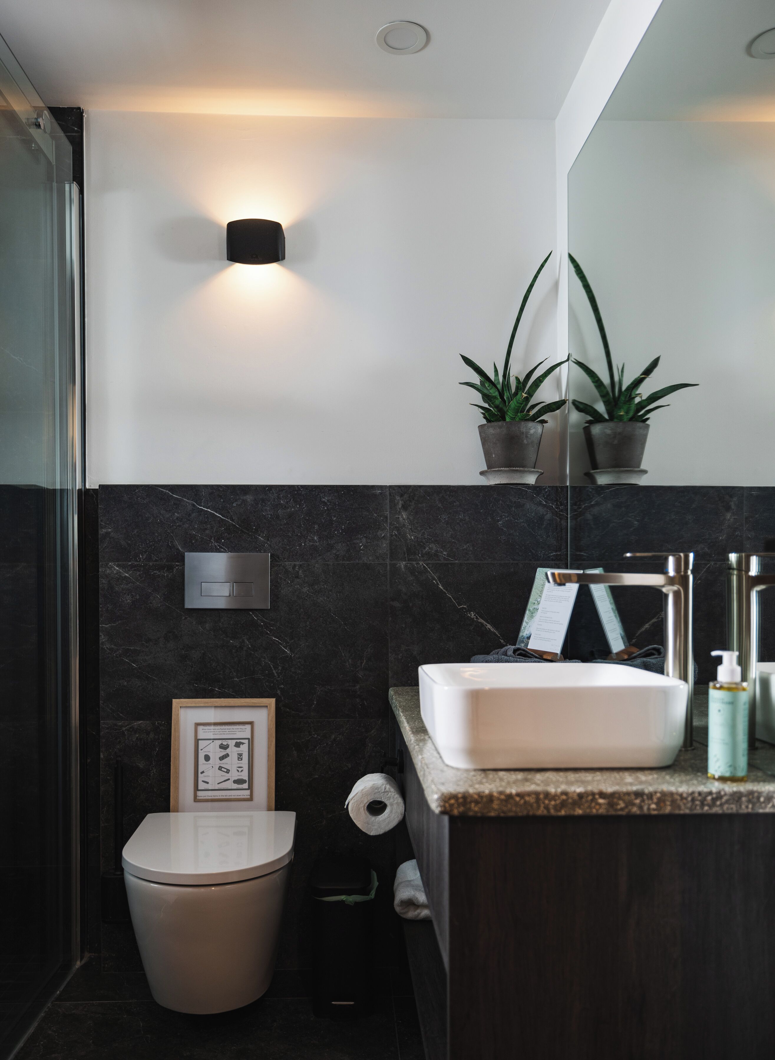 A modern bathroom with a white sink, dark marble walls, a toilet, and two potted plants on a shelf.