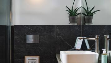 A modern bathroom with a white sink, dark marble walls, a toilet, and two potted plants on a shelf.