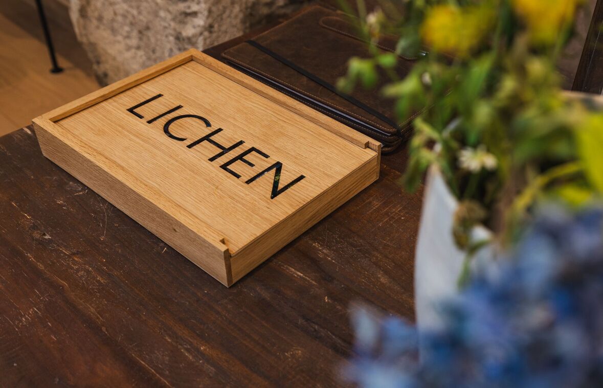A wooden box with the word 'LICHEN' on it, placed on a wooden surface next to a black leather item.