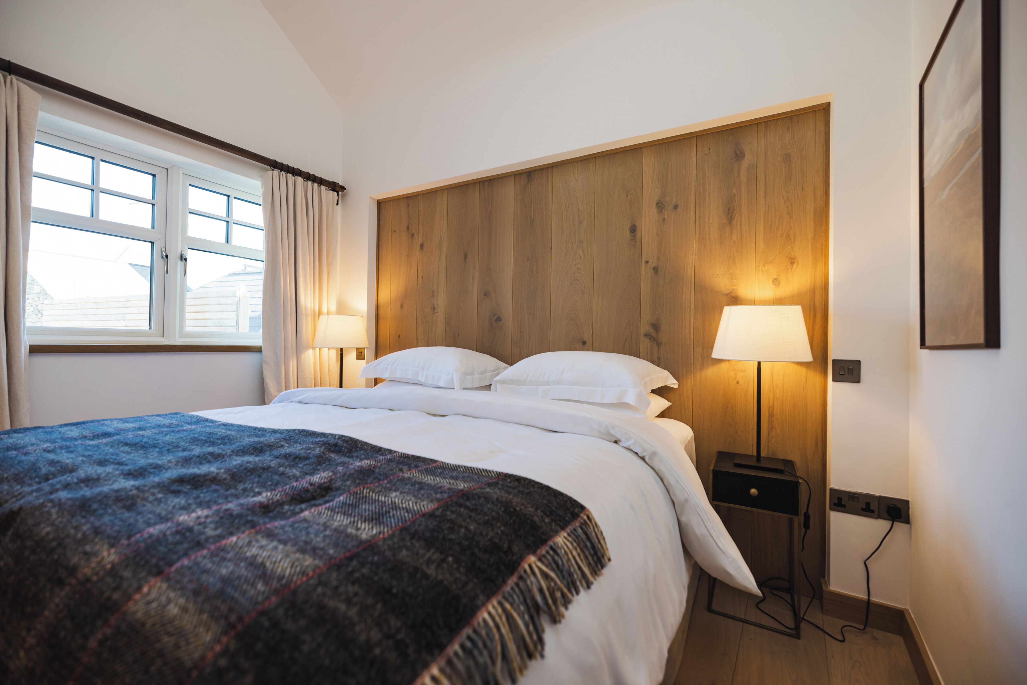 Una habitación de hotel con cama, dos lámparas de noche, cabecero de madera y ventana con cortinas.