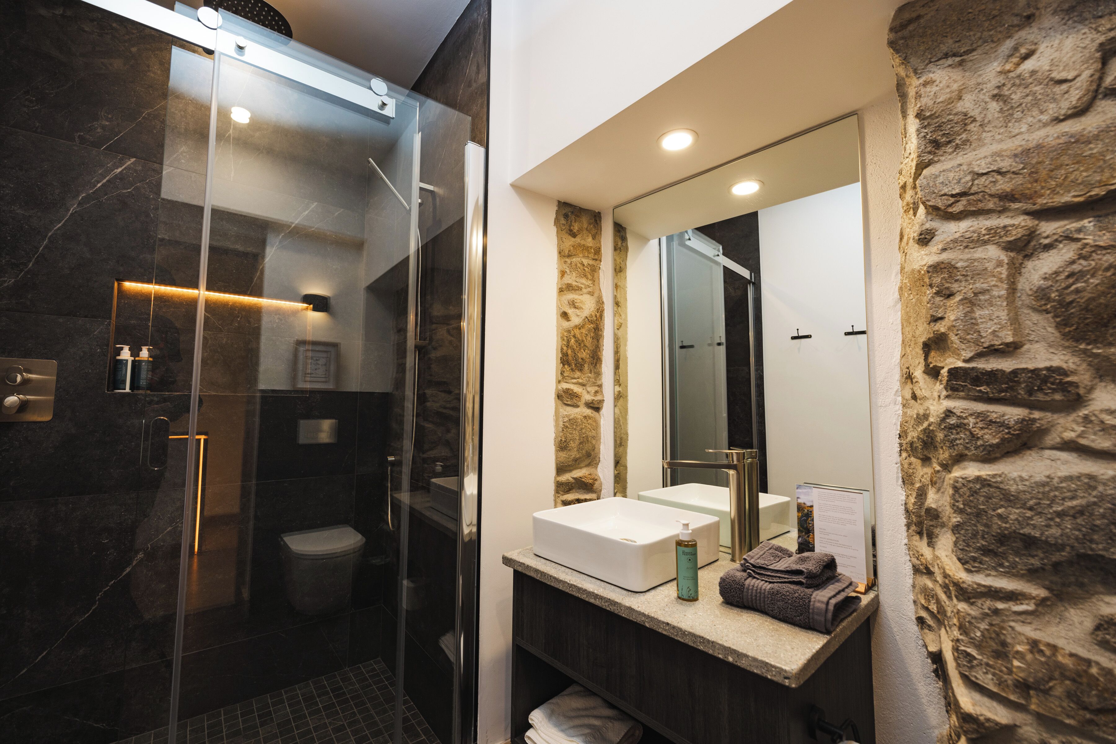 Une salle de bain moderne dotée d’une douche à parois de verre, d’un mur d’accent en pierre et d’un meuble-lavabo avec un miroir.