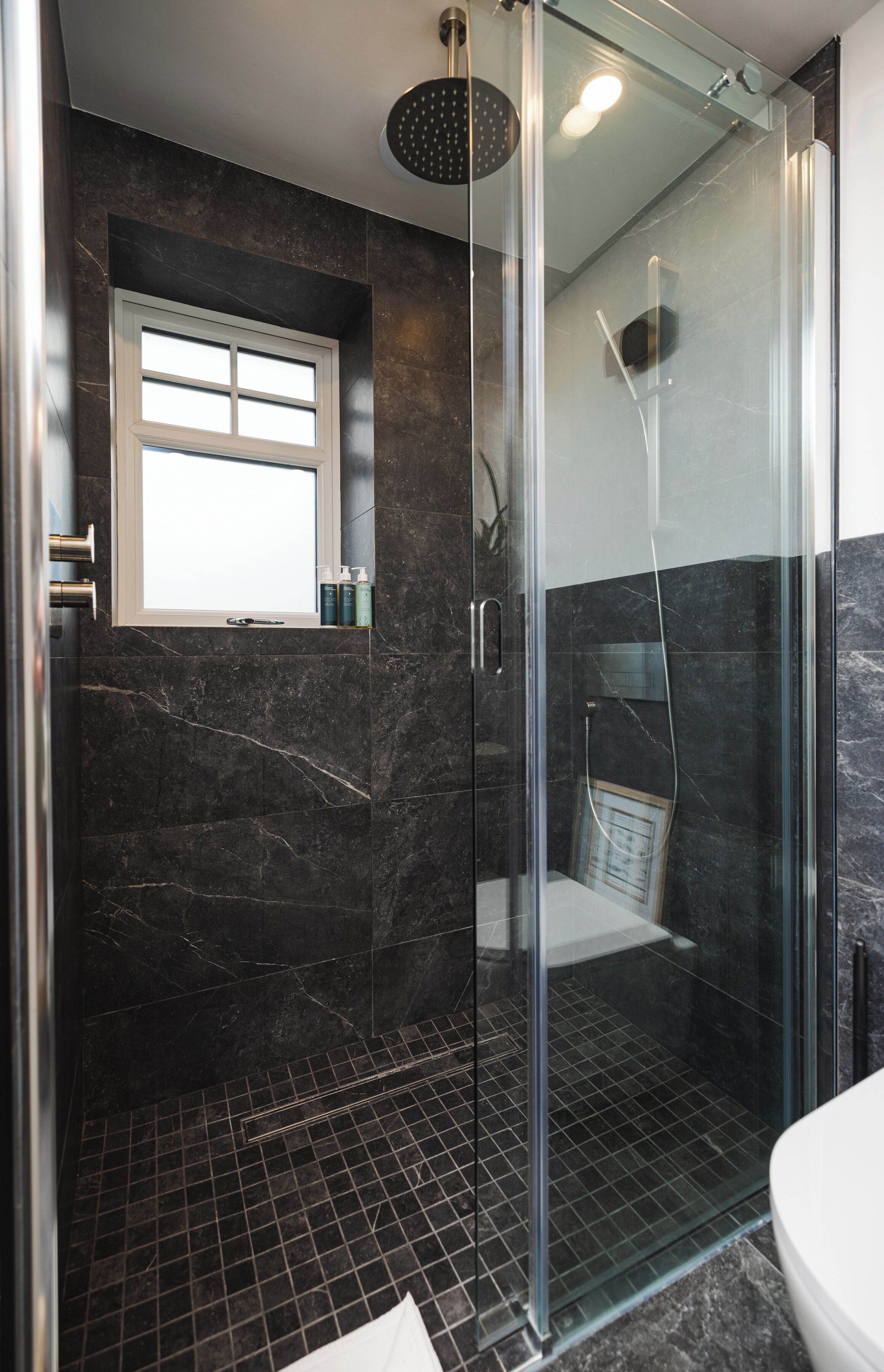 A modern bathroom with a glass-enclosed shower, dark marble walls, and a small window.