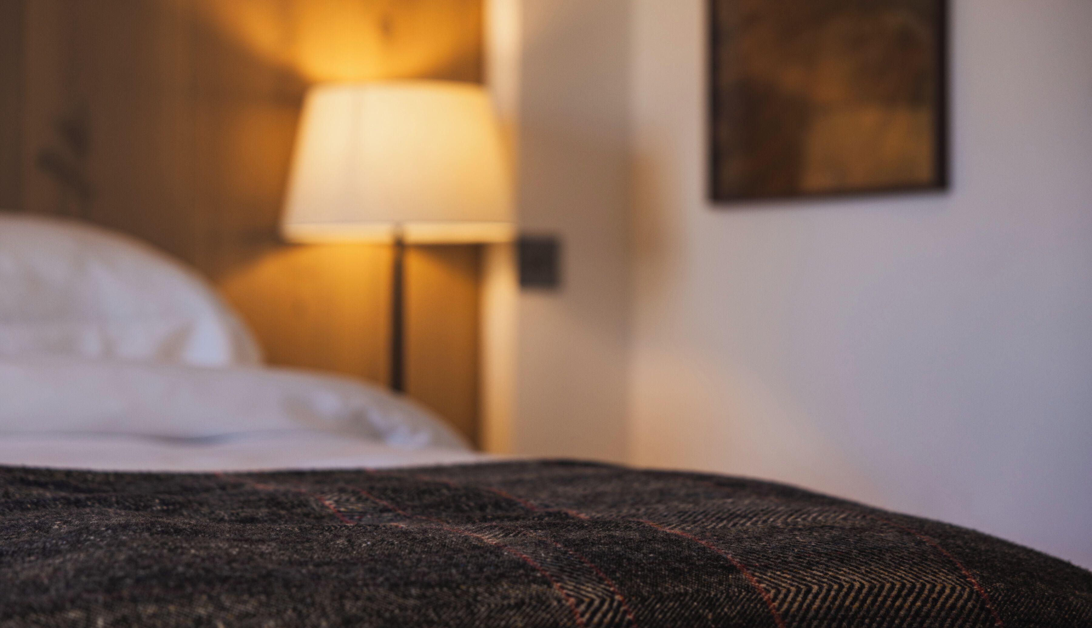 A bed with a dark patterned blanket, a white pillow, and a lamp with a white shade on a bedside table.