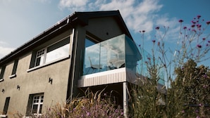 A modern two-story house with a large glass balcony, surrounded by natural vegetation. - Within the Village (Roundstone)