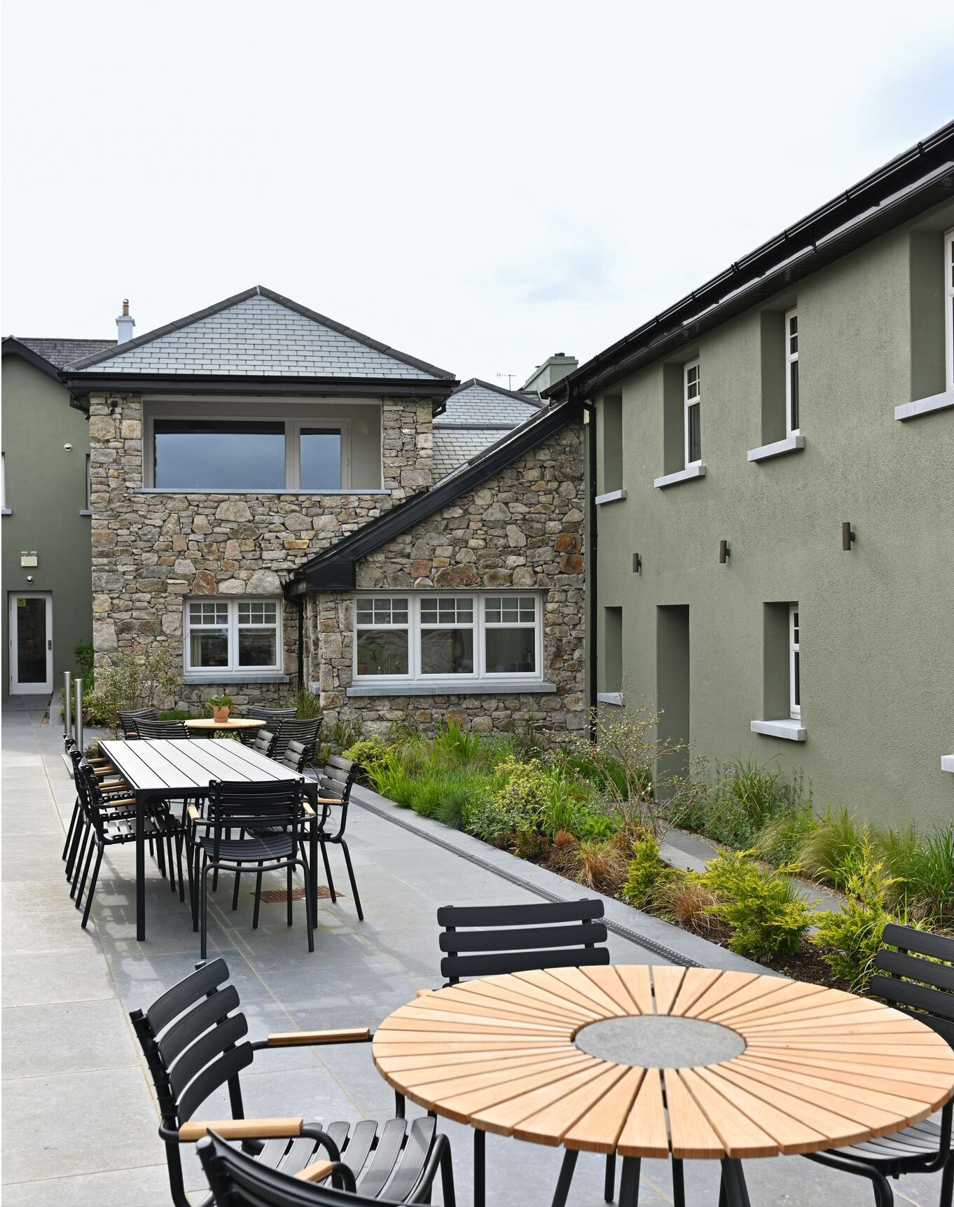 A courtyard with a stone building, a wooden table, and several chairs.