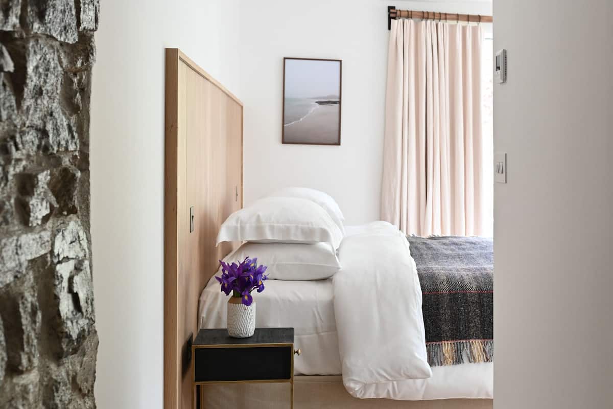 A bedroom with a bed, a nightstand with a vase of flowers, a wooden wardrobe, a framed picture on the wall, and a window with curtains.