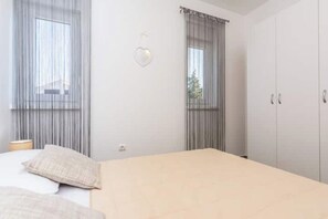 A bedroom with a bed, two windows with curtains, a bedside table, and a wardrobe. - Pasko apartments 3 (Novalja)