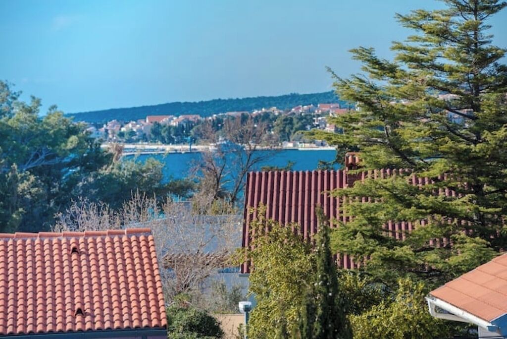 A coastal town with terracotta-roofed buildings, a body of water, and a hill with houses in the background.