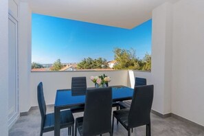 Uma varanda com uma mesa azul e quatro cadeiras, um vaso com flores e uma vista clara de edifícios e árvores ao fundo.